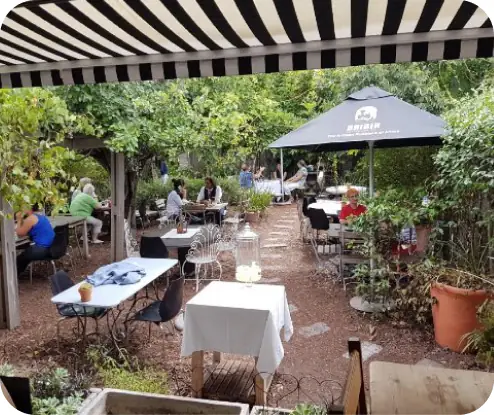 Shaded garden café seating with tables, patrons under striped awning and umbrella
