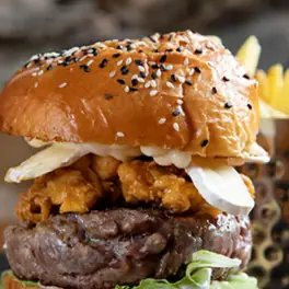 Stacked burger with sesame-topped bun, fried chicken, beef patty and mayo