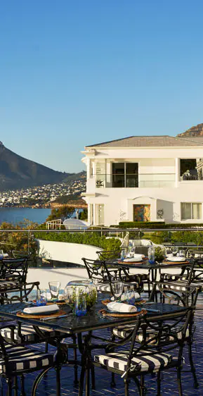 Outdoor dining terrace with set tables overlooking white coastal villa, bay and mountains