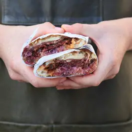 Hands holding a halved wrap filled with purple cabbage slaw and grilled meat